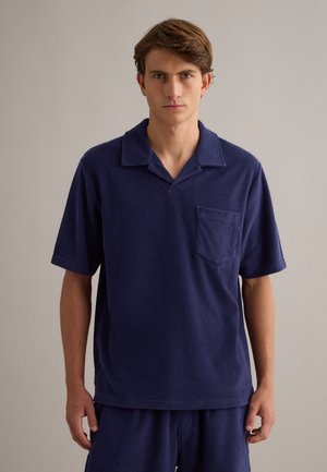 Jeune homme aux cheveux courts et bruns porte un pull en maille bleu marine à manches courtes avec col et poche poitrine, debout devant un fond uni.