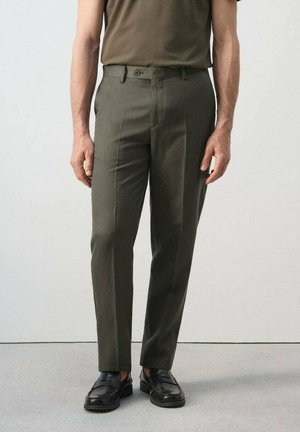 Mann trägt olivgrüne, maßgeschneiderte Hose und schwarze Leder-Loafer, steht vor schlichtem hellem Hintergrund.