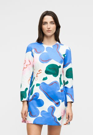 Žena oblečená v krátkych šatách s dlhým rukávom, bieleho základu a veľkými abstraktnými modrými, zelenými a ružovými kvetinovými vzormi na jednofarebnom pozadí.