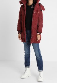 Burgunderroter Steppmantel mit Kapuze, diagonalem Steppmuster, Frontreißverschluss und Druckknöpfen. Kombiniert mit einem schwarzen Pullover und blauen Jeans.