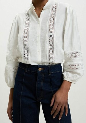 Blusa blanca de manga larga con detalles de encaje en el frente y las mangas combinada con vaqueros azul oscuro de cintura alta en una persona de pie.
