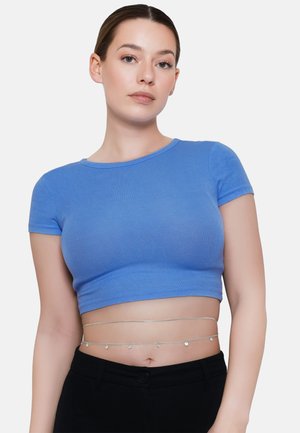 Młoda kobieta w niebieskim krótkim t-shircie, czarnych spodniach i warstwowych srebrnych łańcuszkach na talii na białym tle.