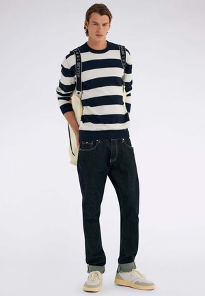 Pull rayé bleu marine et blanc avec un col rond, associé à un jean en denim foncé. Le mannequin porte des baskets claires, montrant une tenue décontractée.