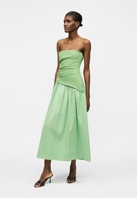 Γυναίκα που στέκεται φορώντας ένα πράσινο τοπ χωρίς τιράντες με υφή και ασύμμετρο τελείωμα και μακριή φούστα που ταιριάζει, σε συνδυασμό με μαύρα ανοιχτά παπούτσια με τακούνι.