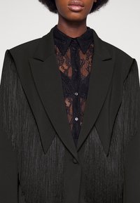 Blazer noir avec détails frangés, doté d'un col cranté et d'une chemise en dentelle en dessous, présentant un design boutonné.