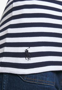 Marinblå och vit randig ribbad tyg, med en liten broderad logotyp av en häst och ryttare. I kombination med mörk denimjeans.