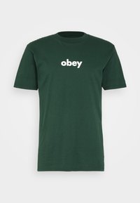 T-shirt en coton vert avec un col rond et des manches courtes, présentant un logo texte "obey" blanc centré sur le devant.