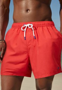 Röda badshorts med elastisk midja, vit dragsko med blå spetsar och en liten blå logotyp på vänster sida. Slät tygtextur.