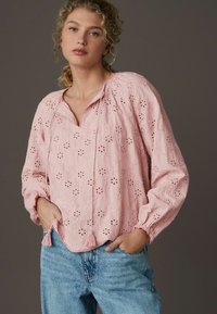 Blusa di tessuto rosa con ricami floreali, scollo a v e maniche lunghe. Include frange all'orlo e una vestibilità rilassata. Indossata con jeans blu.