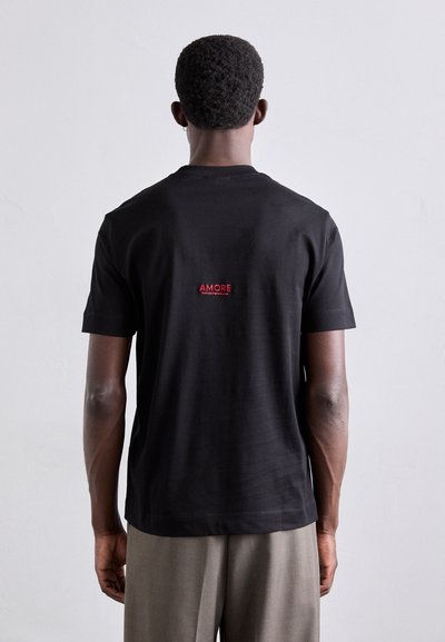 T-shirt noir à manches courtes avec un col rond et un texte "AMORE" rouge dans le haut du dos ; fabriqué en coton doux avec une texture lisse.