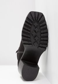 Semelle de botte noire avec des crampons profonds et des motifs en forme de croix, inclinée contre un fond blanc d'angle.