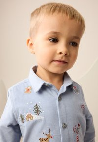 Hellblaues Cordhemd mit gestickten Motiven, darunter ein Lebkuchenmann, ein Rentier, Bäume und Zuckerstangen. Hemdkragen mit Knöpfen.