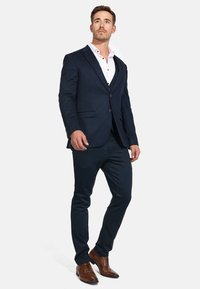 Marineblazer und schmal geschnittene Hose aus strukturiertem Stoff, kombiniert mit einem weißen Hemd. Braune Schnürschuhe mit Durchbrüchen.