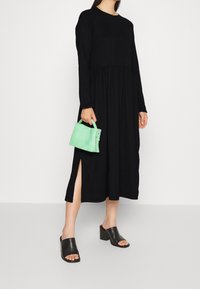 Mujer vistiendo un largo vestido negro con abertura lateral, mules negros de tacón alto y sosteniendo un pequeño bolso verde frente a un fondo blanco liso.