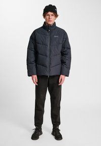 Svart vadderad jacka med hög krage och dragkedja, med en quiltad design. Kombinerad med svarta byxor och sneakers.