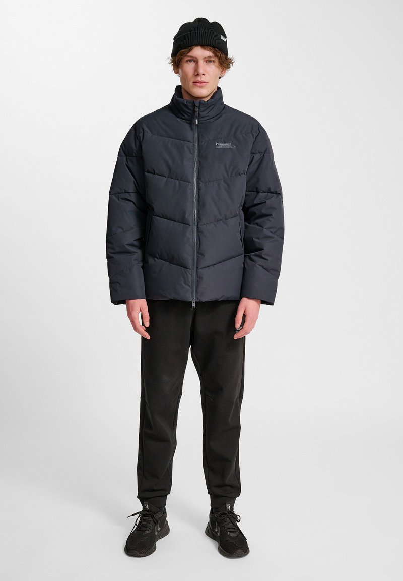 Svart vadderad jacka med hög krage och dragkedja, med en quiltad design. Kombinerad med svarta byxor och sneakers.