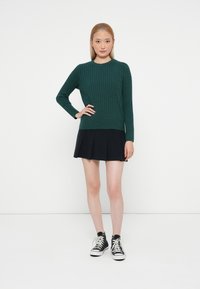 Pull en maille côtelée vert avec des manches longues, associé à une jupe plissée noire et des baskets montantes noires. Silhouette simple et ajustée.
