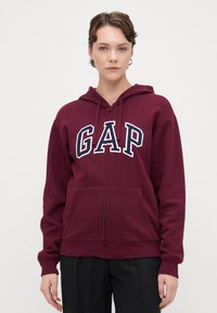 Sudadera con capucha de color burdeos y cremallera, con un logotipo blanco de "GAP" en el pecho, de textura suave y con dos bolsillos frontales.