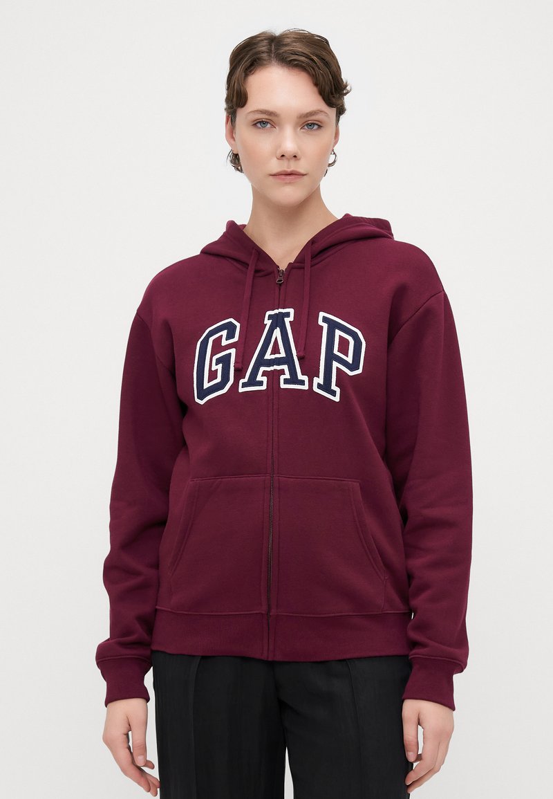 Sudadera con capucha de color burdeos y cremallera, con un logotipo blanco de "GAP" en el pecho, de textura suave y con dos bolsillos frontales.