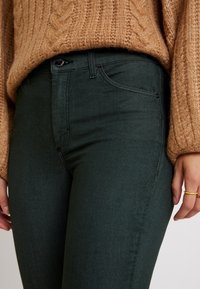 Jeans de mezclilla verdes con un ajuste slim, que presentan un bolsillo frontal, cierre de botón y un desvanecimiento sutil, combinados con un suéter de punto grueso marrón.