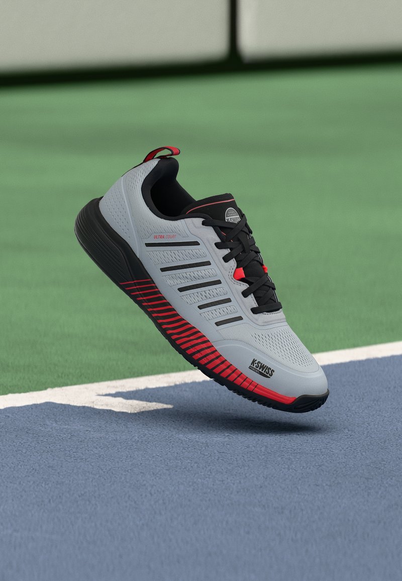Ανοιχτό γκρι παπούτσι τένις K-Swiss Ultra Court με μαύρες και κόκκινες λεπτομέρειες, να αιωρείται πάνω από την επιφάνεια ενός γηπέδου τένις με μπλε και πράσινες αποχρώσεις.