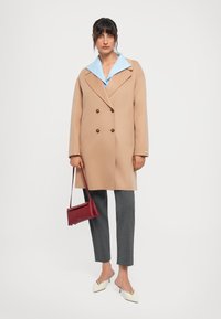 Donna che indossa un cappotto cammello doppiopetto, una camicia blu con colletto, pantaloni grigi alla caviglia, tacchi bianchi e tiene in mano una borsa rossa.