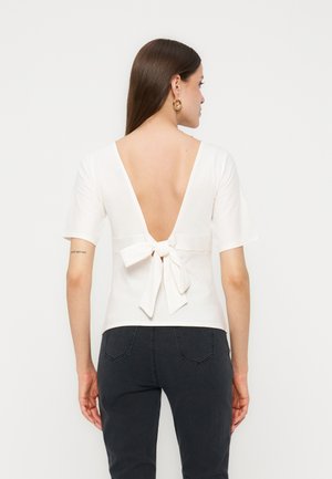 Kvinde med langt brunt hår iført en hvid, kortærmet top med dyb V-ryg bundet med en sløjfe, kombineret med mørke jeans.