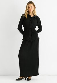 Black pleated maxi dress featuring a button-up front, a tie waist, long sleeves, and scalloped hem. Soft fabric with a smooth texture.