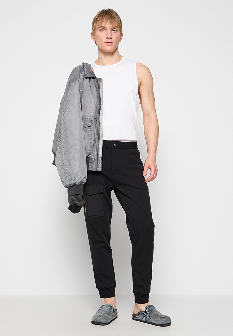 Szara kurtka bomber, biała koszulka bez rękawów, czarne zwężane spodnie cargo z bocznymi kieszeniami oraz szare buty wsuwane o teksturowanej powierzchni.