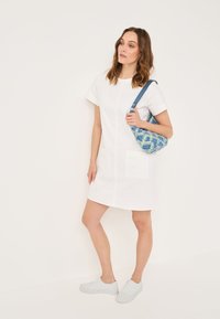 Weißes kurzes Kleid mit kurzen Ärmeln und zwei großen Vordertaschen. Das Modell trägt eine Schultertasche mit blauem und grünem Muster. Weiße Sneaker runden das Outfit ab.