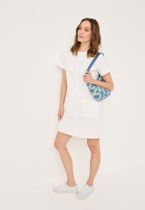 Weißes kurzes Kleid mit kurzen Ärmeln und zwei großen Vordertaschen. Das Modell trägt eine Schultertasche mit blauem und grünem Muster. Weiße Sneaker runden das Outfit ab.