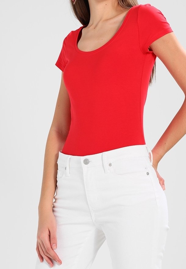 Femme portant un haut rouge ajusté à manches courtes et un jean blanc taille haute avec boutons et poches, debout devant un fond uni.