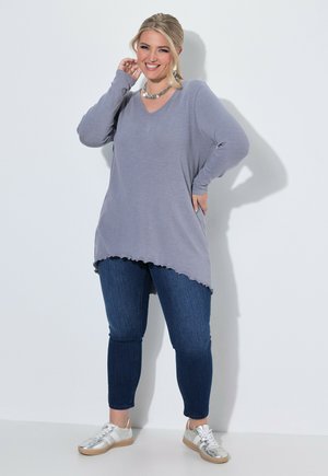 Lachende vrouw die een losse grijze lange mouw top draagt, donkerblauwe spijkerbroek, zilveren sneakers en een zilveren ketting, staand tegen een witte muur.