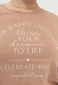 Světle hnědé bavlněné tričko s krátkým rukávem, které má bílý textový design. Text zahrnuje motivační fráze a symbol srdce.