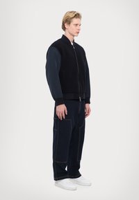 Giubbotto bomber nero con maniche blu navy, abbinato a pantaloni cargo larghi di un blu scuro con cuciture visibili e tasche. Sneakers bianche completano il look.