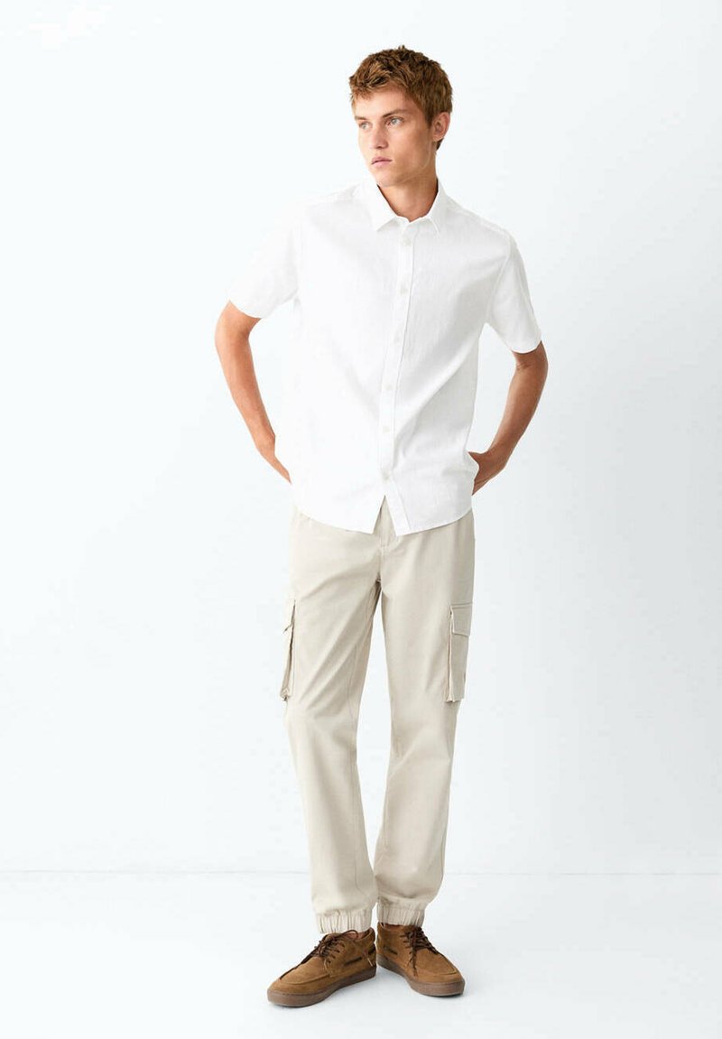 Jeune homme en chemise blanche à manches courtes boutonnée, pantalon cargo beige et chaussures marron, debout les mains sur les hanches regardant vers la droite.
