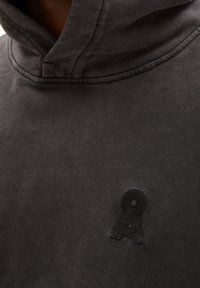 Šedá mikina z měkkého materiálu, s kapucí na šňůrku a černým vyšitým logem na prsou. Texturovaný povrch se šitými švy.