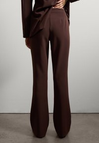 Pantalons brun sur mesure avec un design évasé, présentant une texture lisse et de subtils plis à la taille, fabriqués à partir d'un tissu léger.