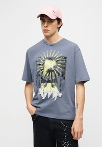 Νέος άνδρας που φοράει γκρι γραφικό t-shirt Puma, ροζ καπέλο Puma και μαύρο παντελόνι με άσπρες κουκίδες, στέκεται με το ένα χέρι στην τσέπη.