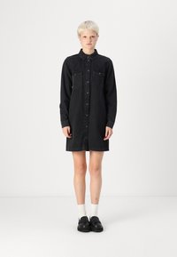 Black denim shirt dress with button closure, collar, and two chest pockets. Long sleeves and knee-length cut. Pair with black shoes and white socks.