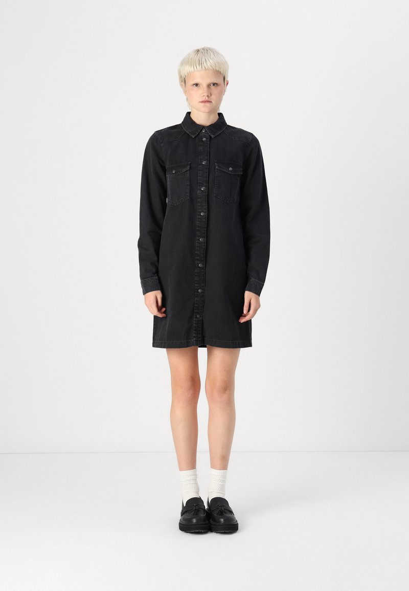 Black denim shirt dress with button closure, collar, and two chest pockets. Long sleeves and knee-length cut. Pair with black shoes and white socks.