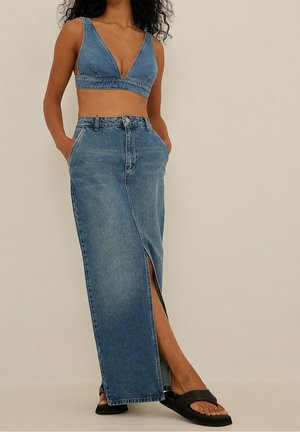 Kobieta ubrana w niebieski krótki top z denimu i pasującą długą spódnicę z rozcięciem z przodu, stojąca z rękami w kieszeniach, w czarnych sandałach.