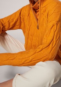 Maglione arancione a maglia con colletto alto e mezza zip. Modello seduto in pantaloni chiari arrotolati, mostrando texture e design.