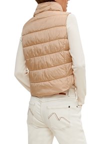 Prošívaná vesta světle béžové barvy s vysokým límcem, s vodorovným prošíváním. Kombinovaná s bílými kalhotami, které zdůrazňují dekorativní švy na kapsách.