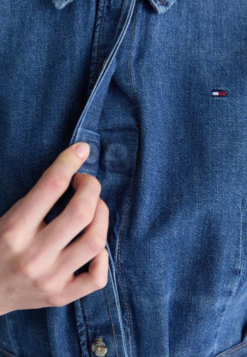 Blue denim button-up shirt with a pointed collar, showing a hand adjusting the placket and a small branded logo stitched in red, white, and blue.