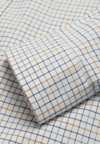 Tela blanca con un patrón de cuadrícula, con líneas azules y naranjas. La textura es suave y el puño de la camisa está doblado con costuras limpias.