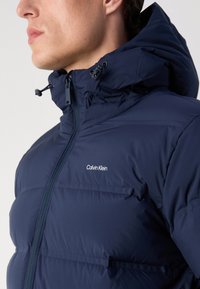 Námornícky pufferovač s vysokým golierom a zipsovým zapínaním. Obsahuje logo Calvin Klein, nastaviteľnú kapucu a horizontálny prešívaný vzor.
