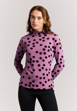 Jeune femme aux cheveux bruns ondulés portant un col roulé violet à grands pois noirs et un pantalon noir, se tenant devant un fond uni.