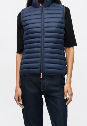 Granatowa pikowana kamizelka bez rękawów z zamkiem z przodu, noszona na czarnym topie z krótkim rękawem i ciemnych dżinsach.