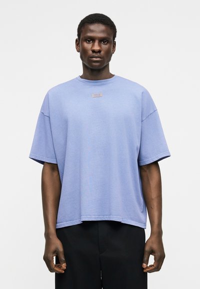 Νεαρός άνδρας με σκουρόχρωμο δέρμα που φοράει ένα φαρδύ ανοιχτό μπλε μπλουζάκι και μαύρο παντελόνι, στέκεται μπροστά σε λευκό απλό φόντο.